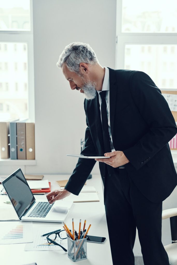 Thoughtful mature man in full suit using laptop and digital tablet while working in modern office